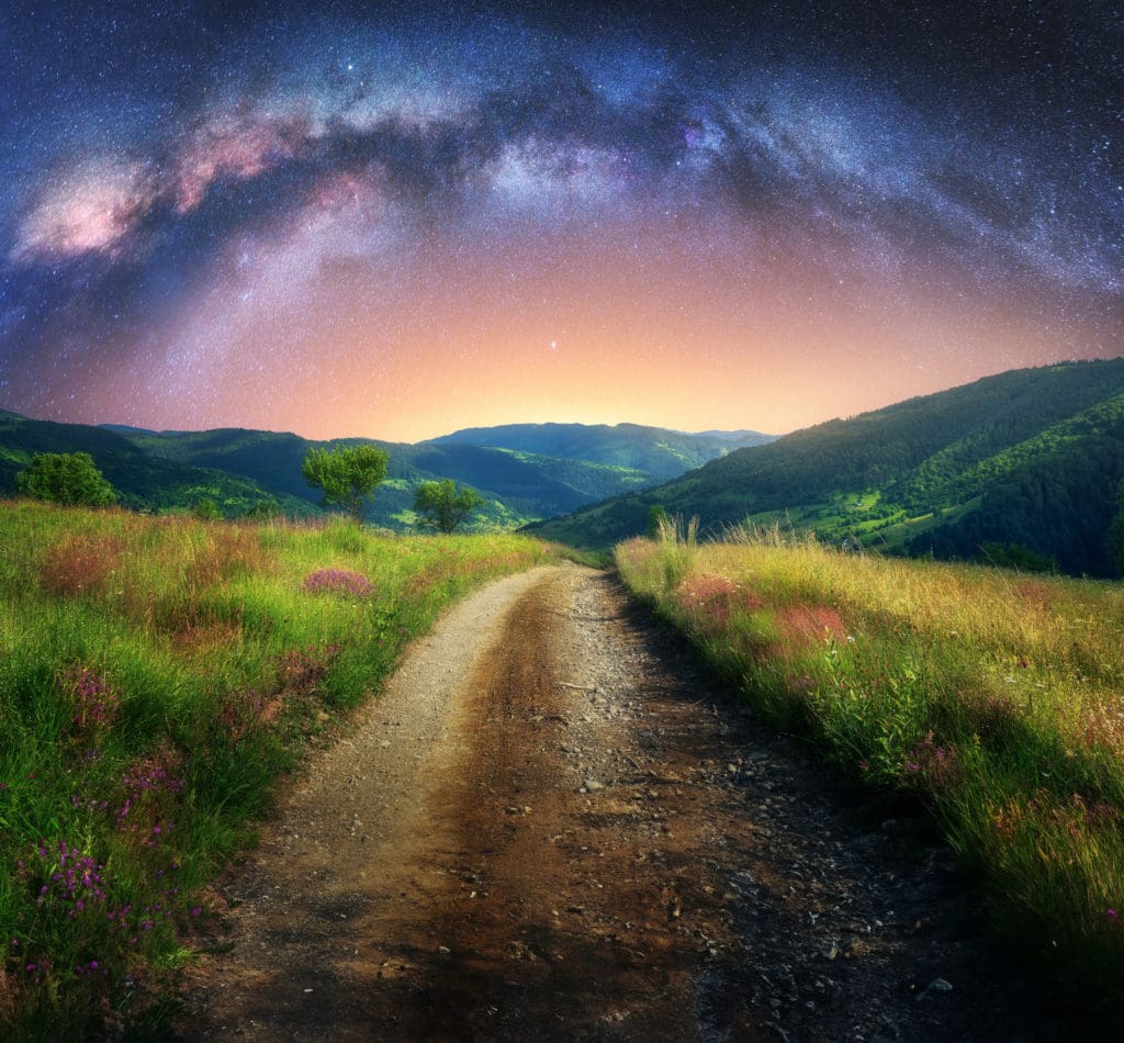 Arched Milky Way over the mountain dirt road in summer. Beautiful night landscape with starry sky, milky way arch, trail in mountain village, hills, green grass and purple flowers. Space and galaxy